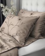 Beige satin pillowcases and quilt with black floral embroidery on a bed near a tufted white headboard, with white flowers in the background.