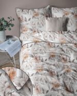 Bed dressed in cream floral duvet and pillows with peach and gray flowers, beside a small table with a plant and open book.