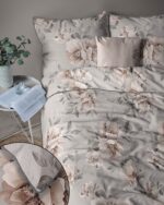 Bed dressed in muted gray floral bedding with matching pillows; a side table holds an open book and a glass beside a potted plant.