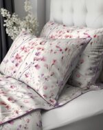 White bedding set with lavender-pink floral print and purple piping on two matching pillows resting on a tufted white headboard.