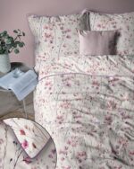 Bedroom scene with a pink-and-purple floral duvet cover and matching pillows on a bed, pink wall in background, and a side table with a plant and an open book nearby.