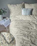 Bed dressed in beige floral duvet and matching pillows, with a blue accent pillow; side table holds an open book, glass, and a plant nearby.